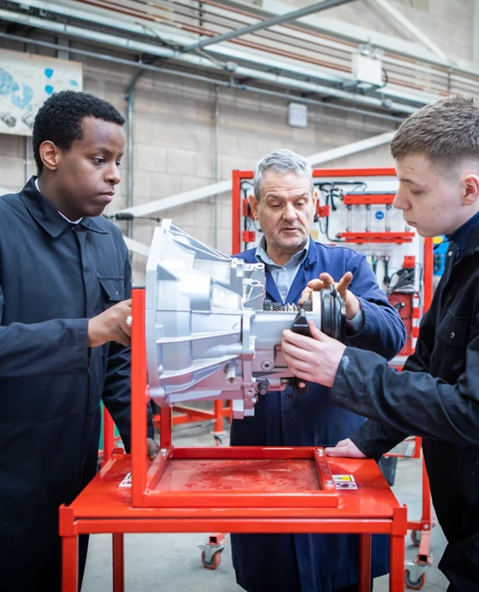 Automotive instructor guiding two students in overalls as they work on a mechanical component in a workshop. Automotive instructor guiding two students in overalls as they work on a mechanical component in a workshop.